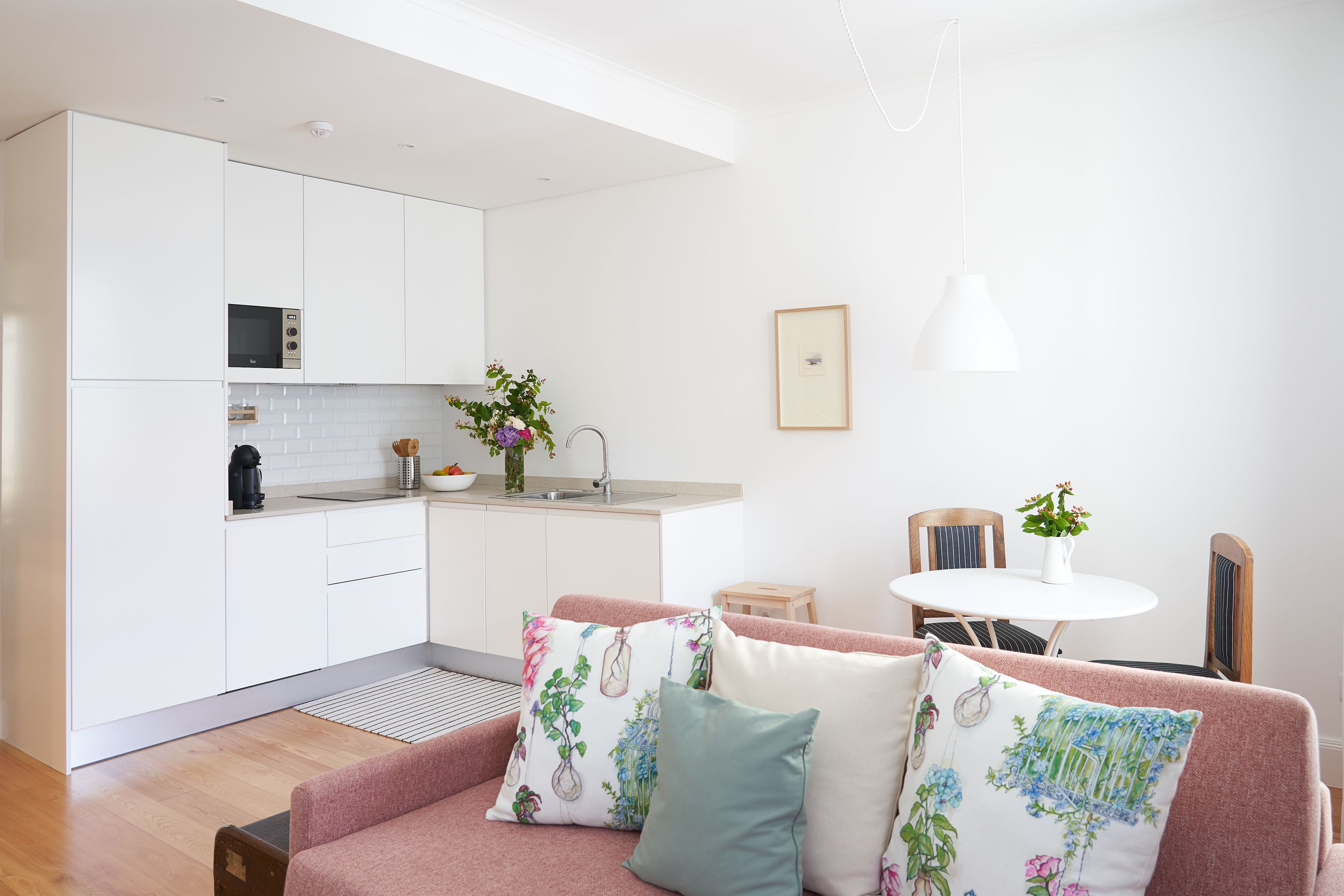 Une cuisine moderne avec des meubles blancs, un four à micro-ondes et un évier. On y trouve un coin repas avec une table et des chaises, ainsi qu’un canapé agrémenté de coussins décoratifs.