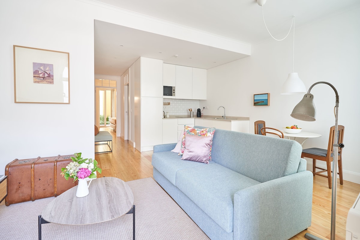 A modern living room with a light blue sofa, a small round table with a vase of flowers, and a vintage suitcase.