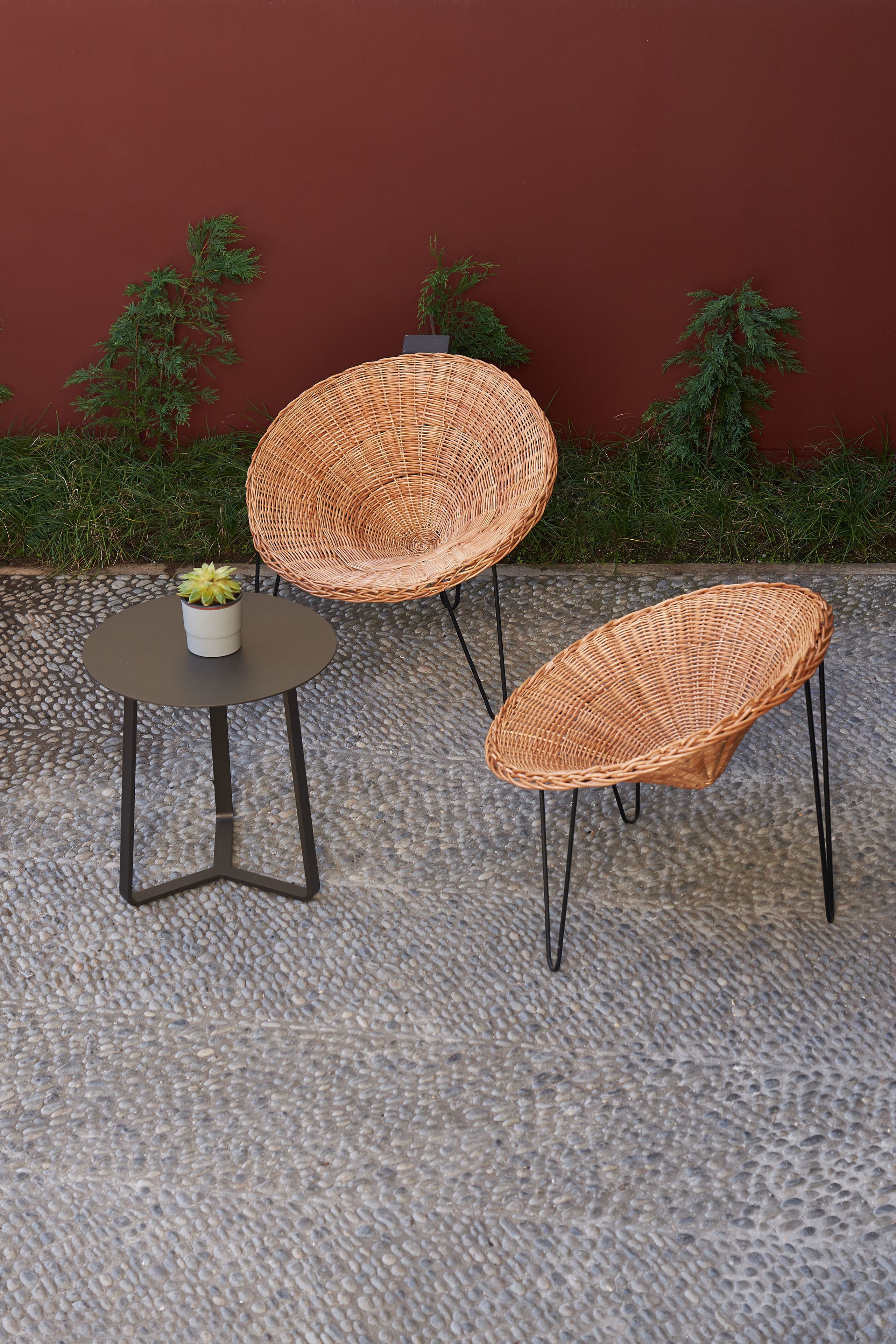 A wicker chair and a small table with a potted plant, set on a textured stone floor against a red wall.
