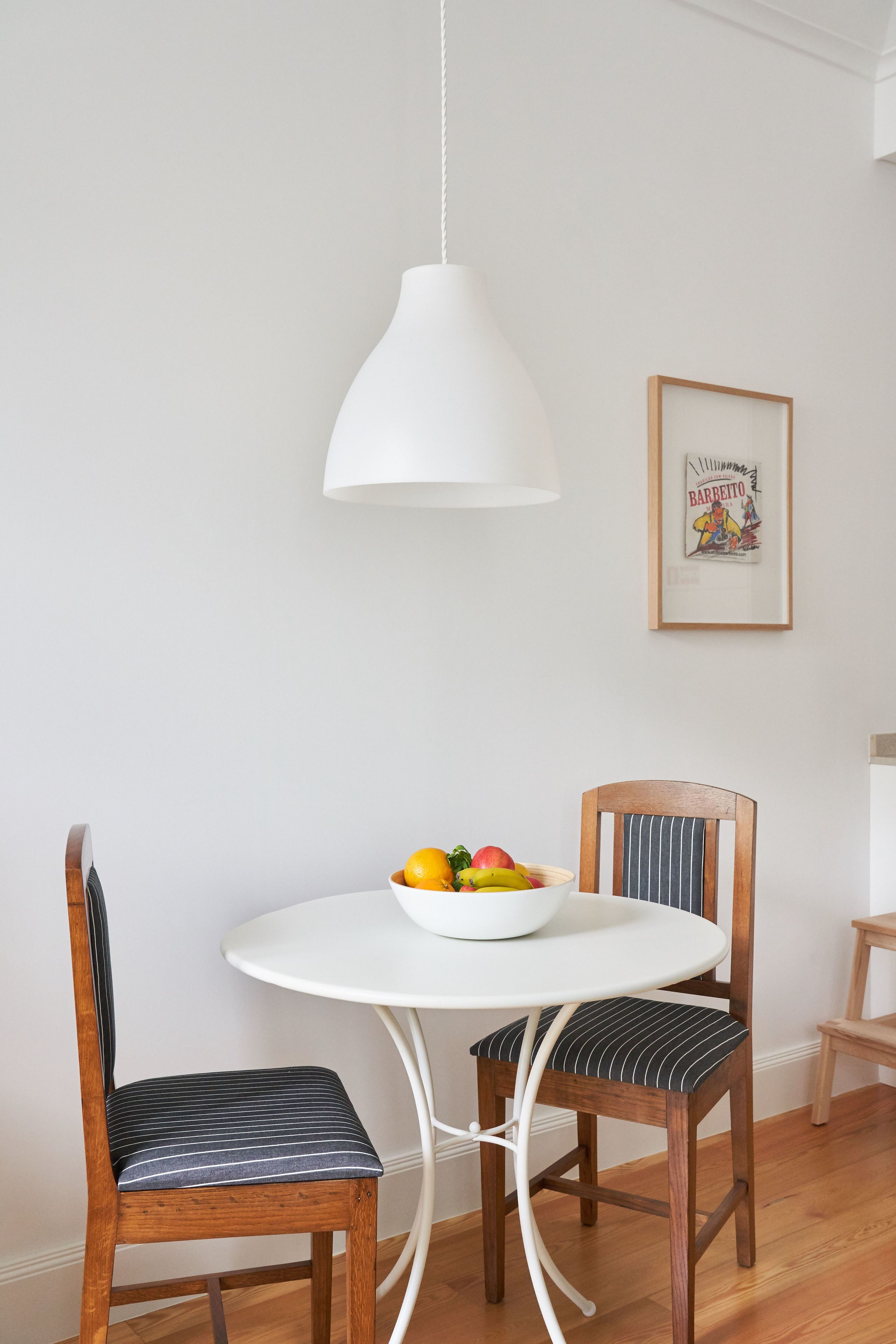 Uma sala de jantar com uma mesa branca redonda, duas cadeiras de madeira com almofadas listradas, uma luminária pendente branca e um quadro na parede.