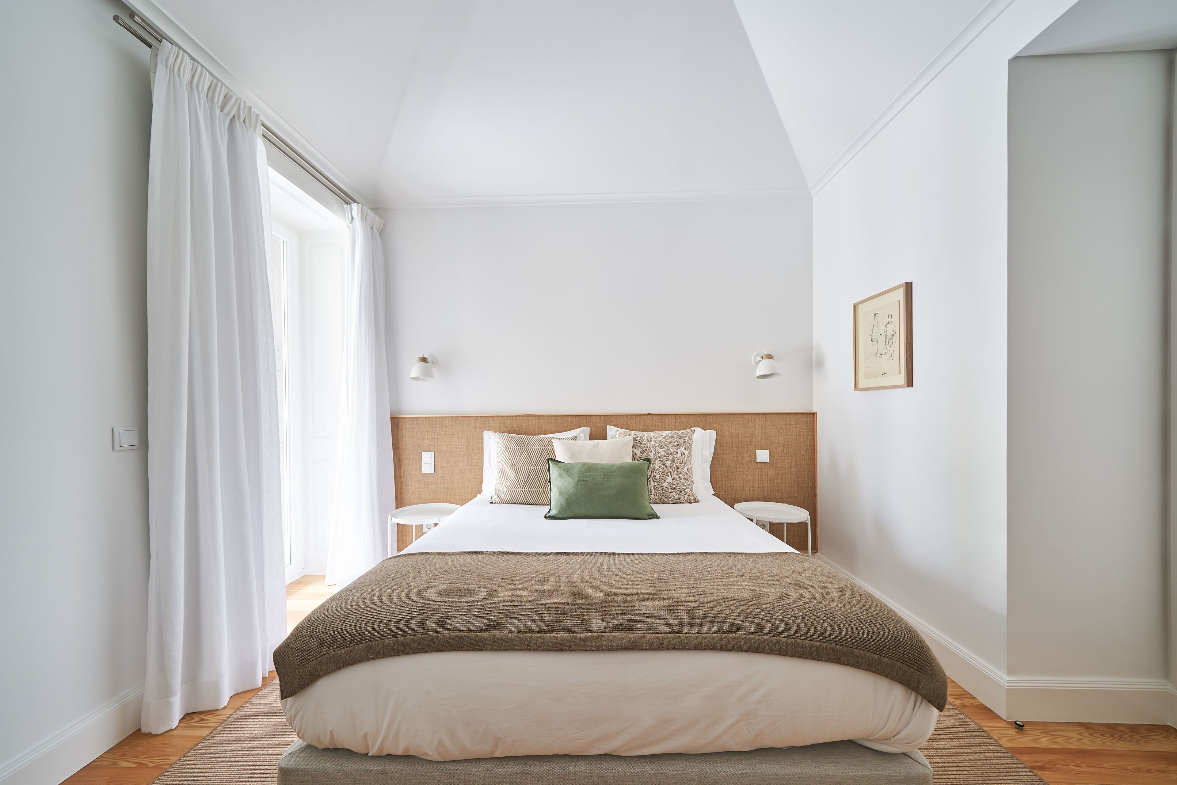 A bedroom with a large bed, a wooden headboard, white walls, and a window with white curtains.