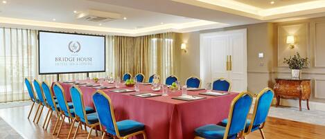 Une salle de conférence avec une table rouge, des chaises bleues et un écran de projection affichant le texte « BRIDGE HOUSE HOTEL, SPA & LEISURE ».