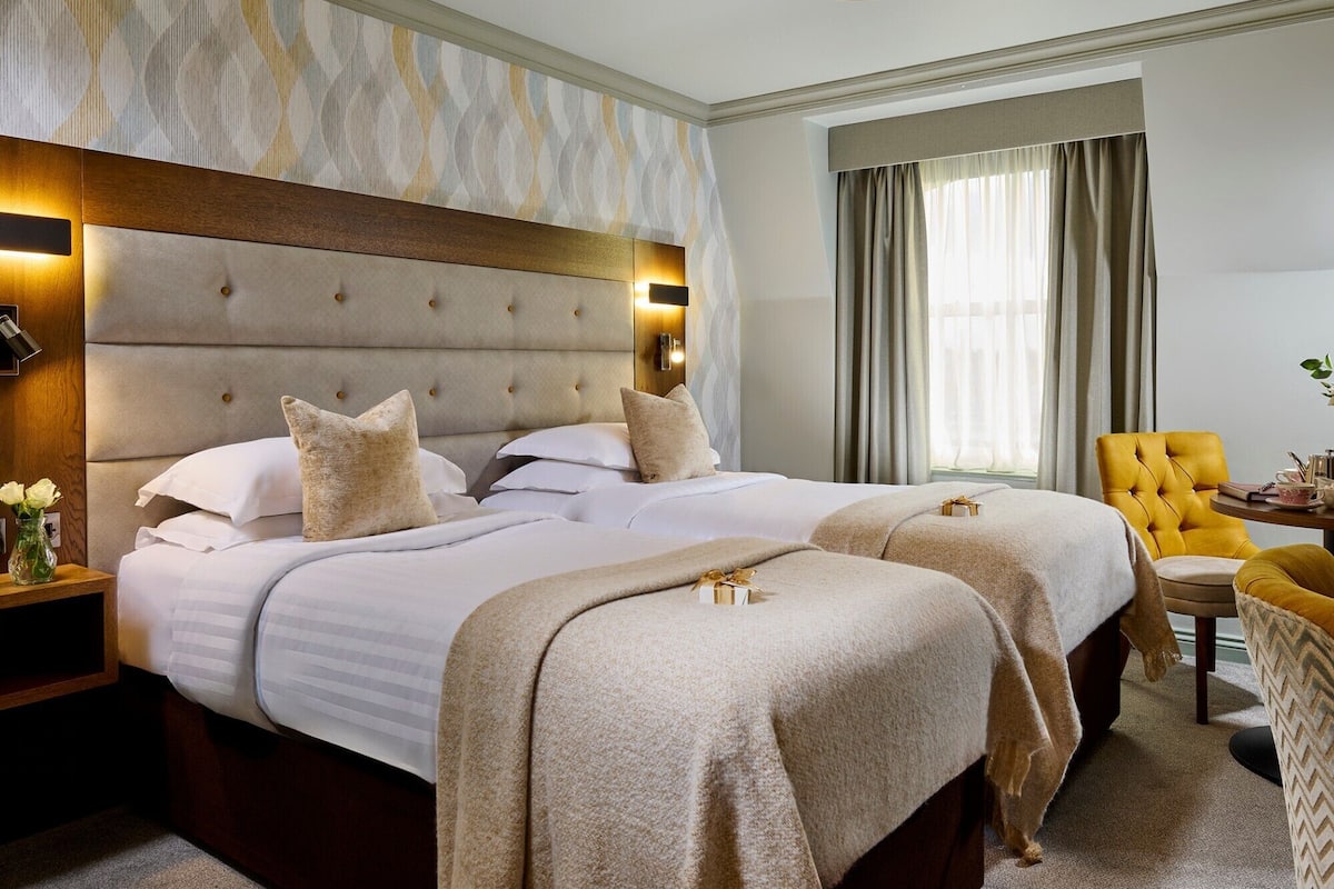 A hotel room with a large bed, a headboard with a light fixture, a window with curtains, and a small table with a chair.