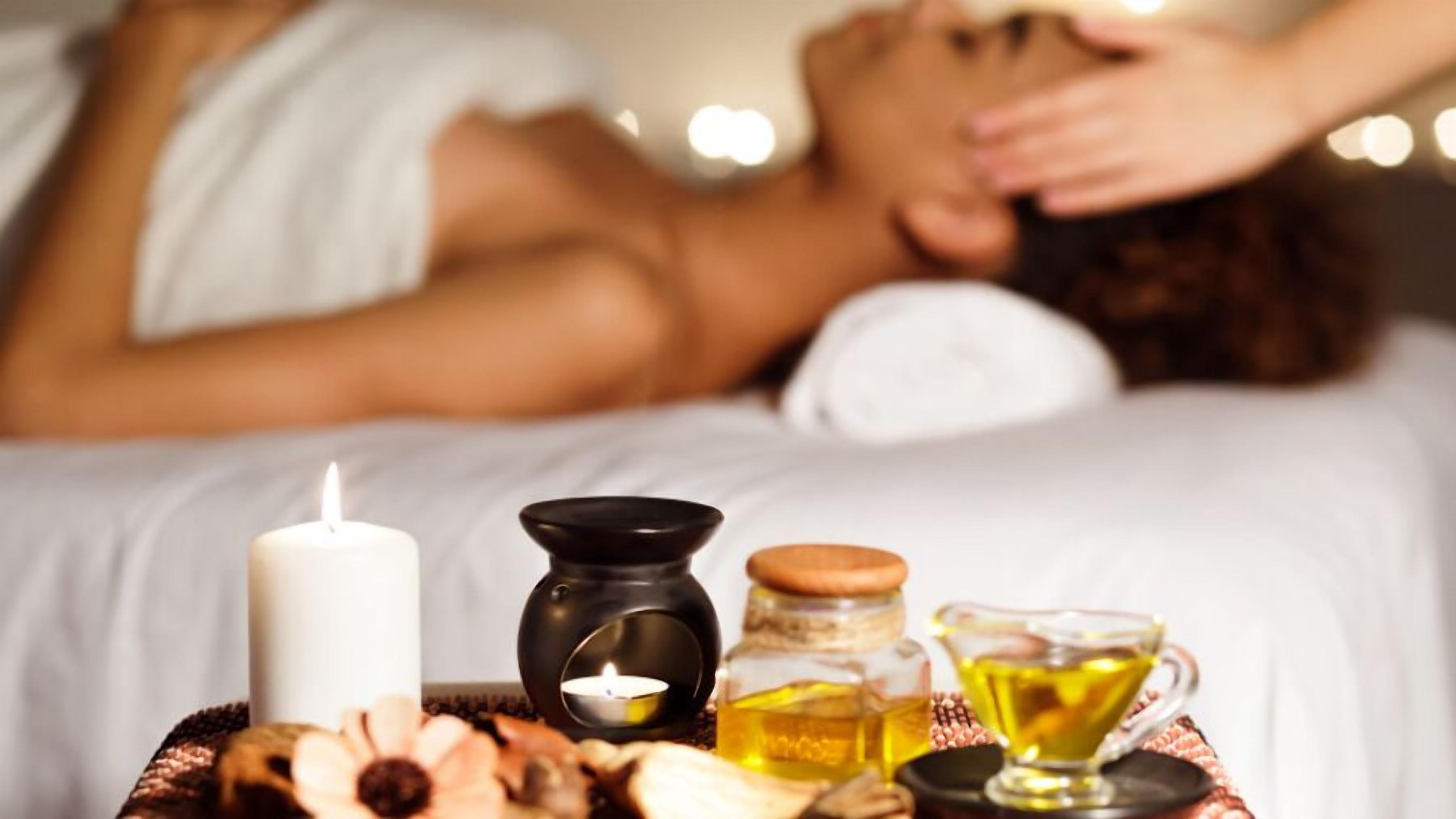 A spa setting with a person receiving a massage, a candle, essential oils, and a glass of oil on a tray.