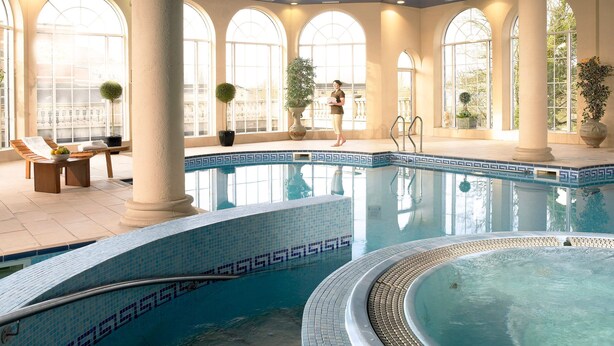 A large indoor swimming pool with a tiled edge, surrounded by columns and large windows. - Bridge House Hotel - Leisure Club & Spa (Tullamore)