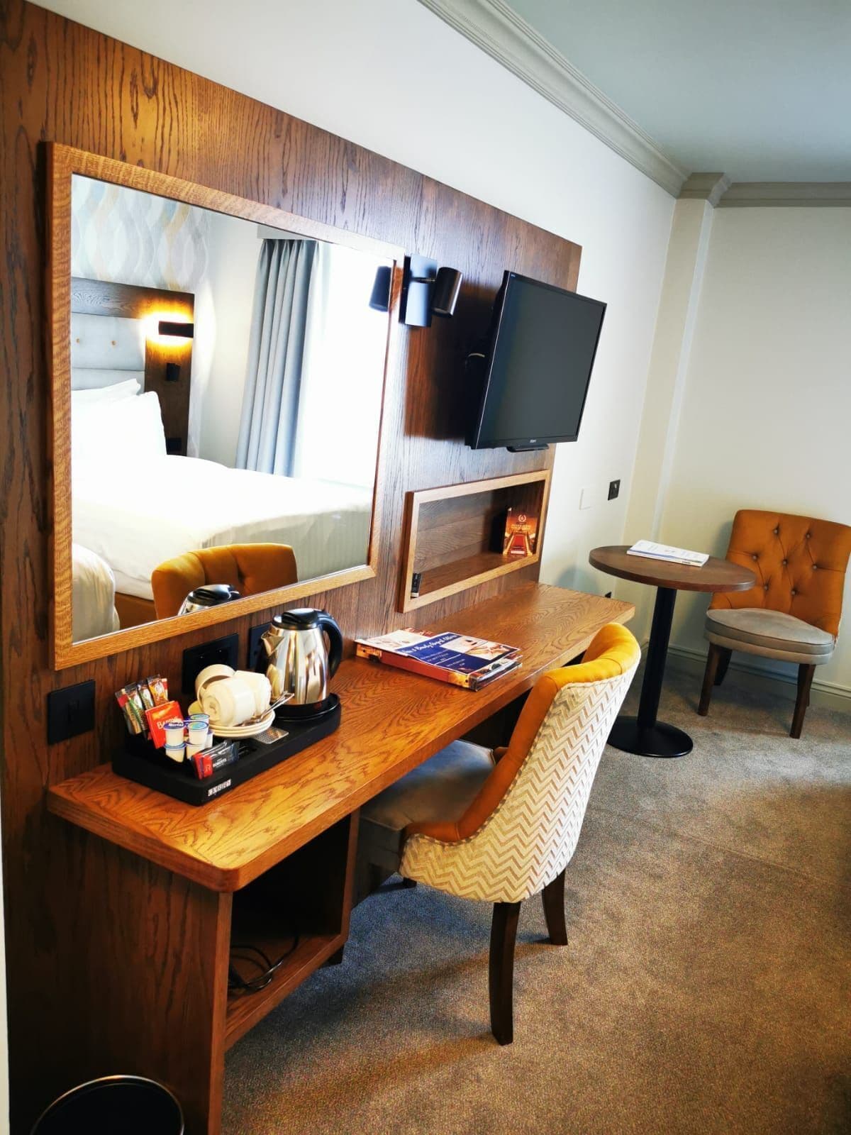 Habitación de hotel con un escritorio de madera, un espejo grande, televisión, una silla y una mesita.