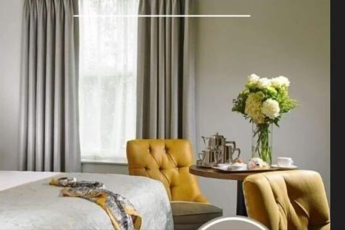 A hotel room with a large bed, a tufted headboard, a yellow armchair, a small round table with a vase of flowers, and a window with curtains.