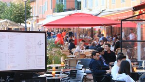 Un café en plein air avec des tables et des chaises, un panneau affichant le menu et un parasol rouge.