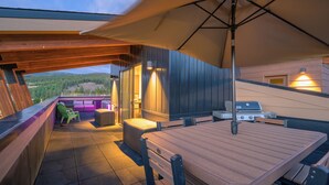 An outdoor patio with a wooden table and chairs, a barbecue grill, and a view of the surrounding landscape.