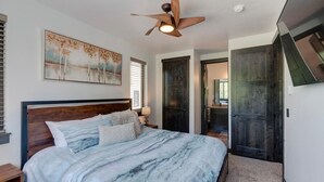 A bedroom with a large bed, a ceiling fan, a painting on the wall, and a view into the bathroom.