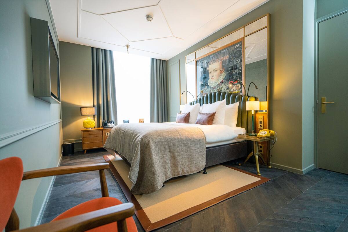 Quarto de hotel moderno com uma cama, escrivaninha, cadeira e uma janela grande com cortinas.