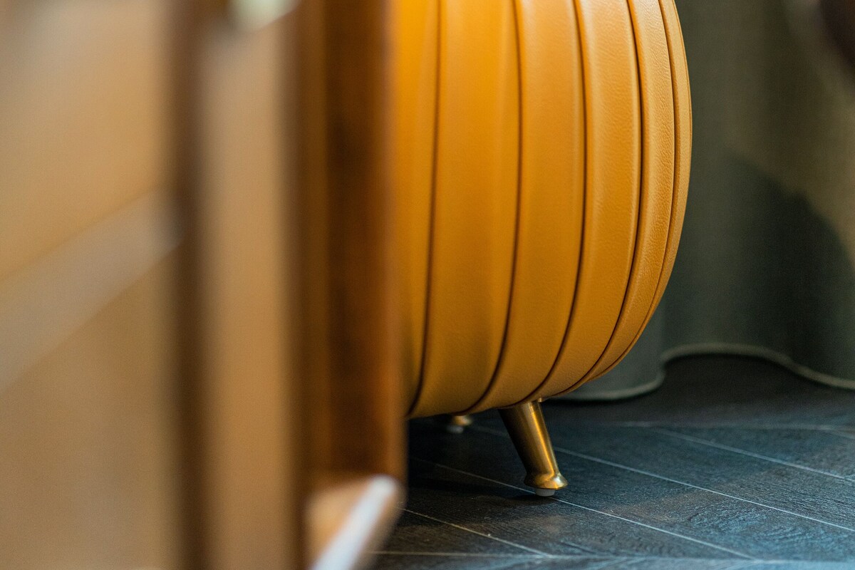 A close-up of a vintage orange leather stool with a curved backrest and metallic legs, positioned in front of a wooden cabinet with a visible handle.
