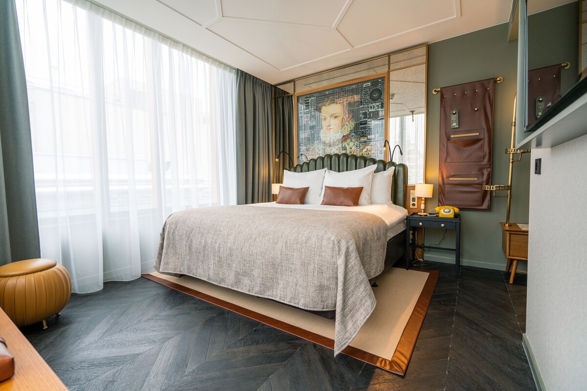 A modern hotel room with a large bed, a desk, a chair, a nightstand, and a large window with curtains.