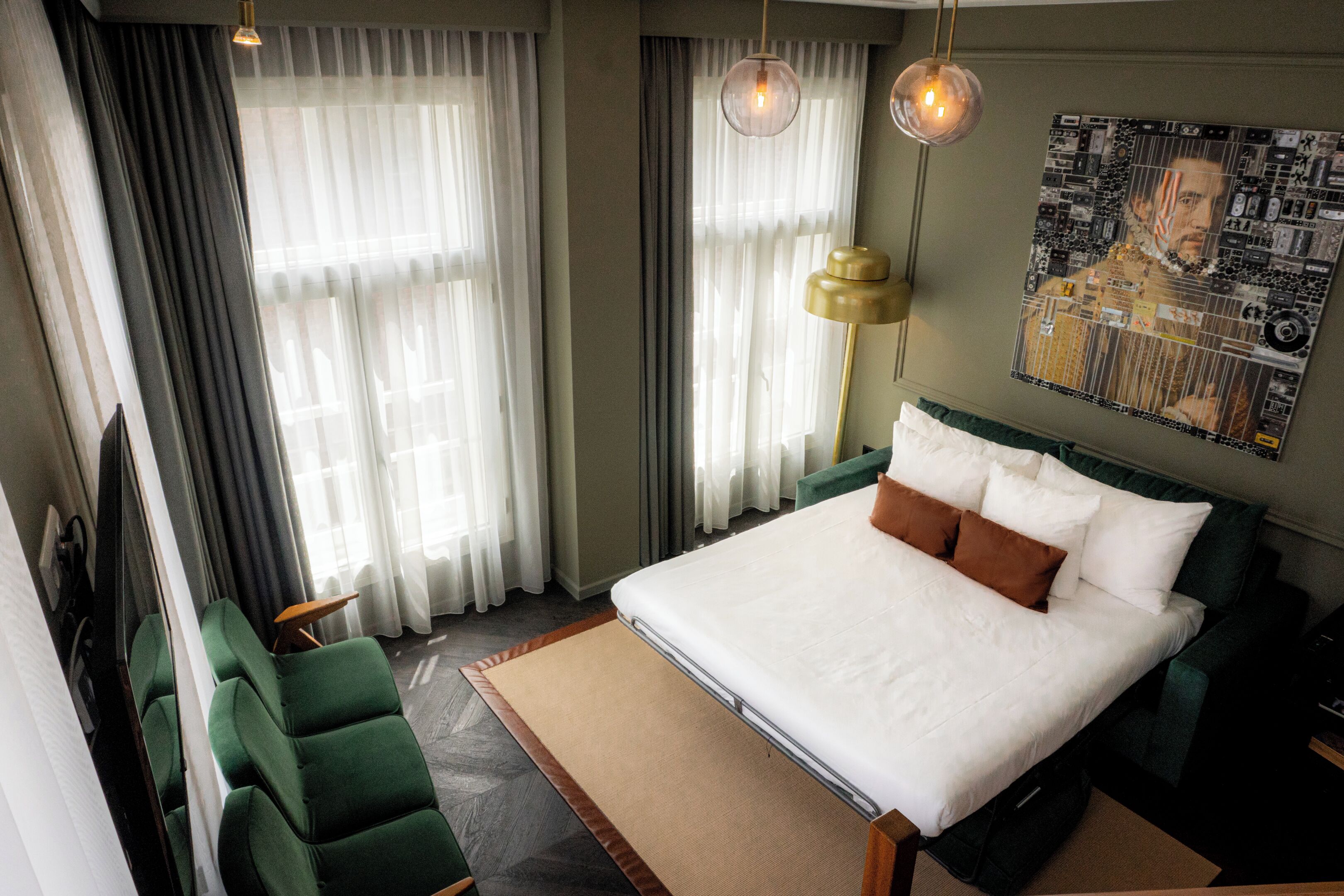 Quarto de hotel moderno com cama grande, poltronas verdes, televisão de tela plana e uma pintura urbana na parede.