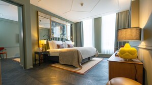 A modern hotel room with a large bed, a bedside table with a lamp, a mirror, and a view of the city through the window. - The Collector (The Hague)