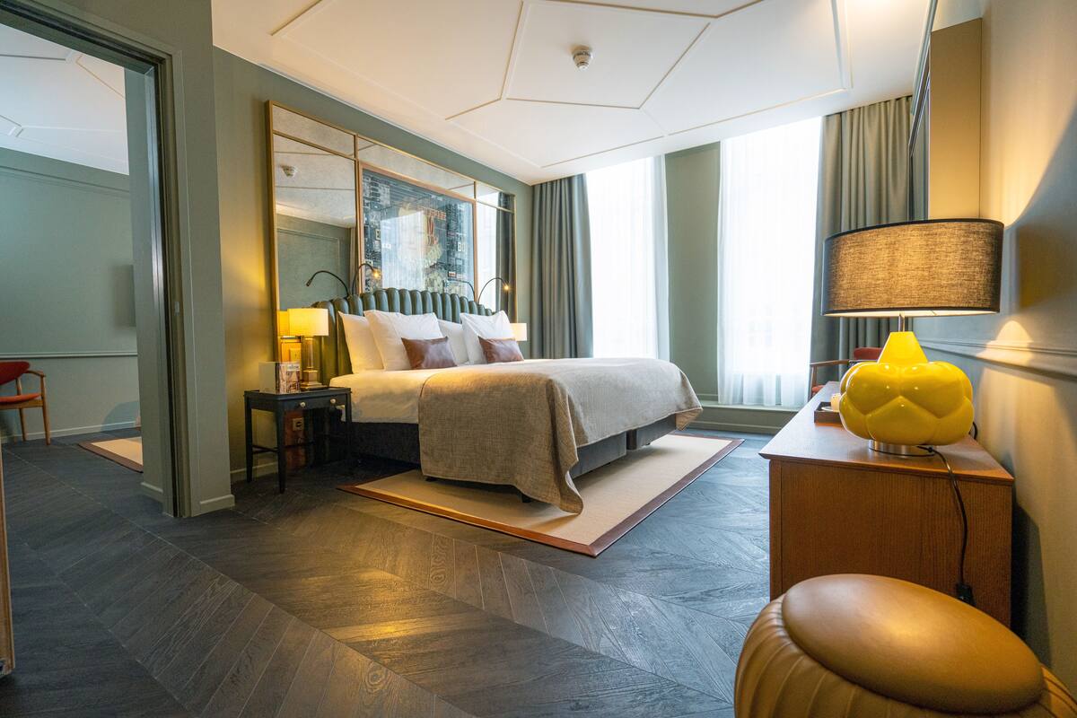 Quarto de hotel moderno com uma cama grande, mesa de cabeceira com abajur, espelho e vista para a cidade pela janela.