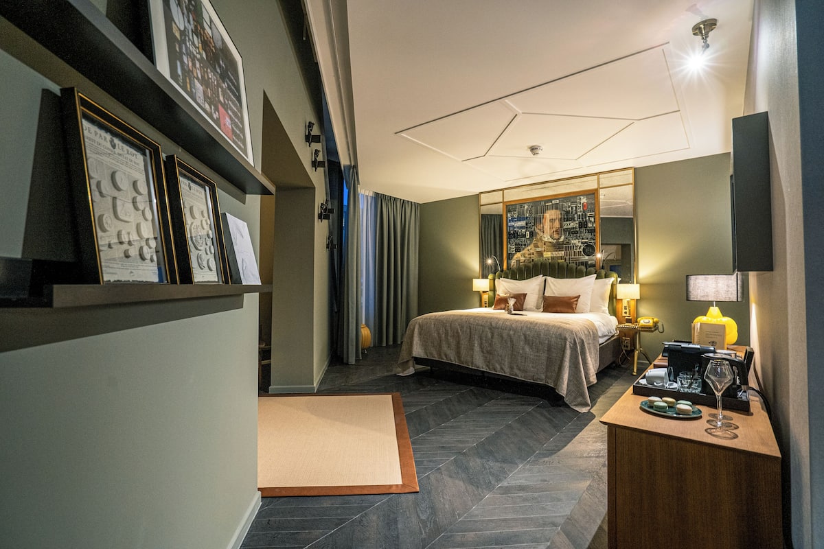 Quarto de hotel com cama, mesa de cabeceira, abajur e quadros na parede.