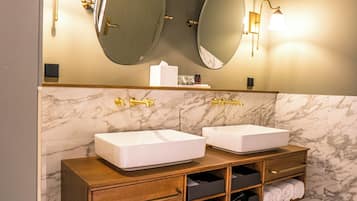 Un baño con dos lavamanos, paredes de mármol y un mueble de tocador de madera con espacio para toallas.