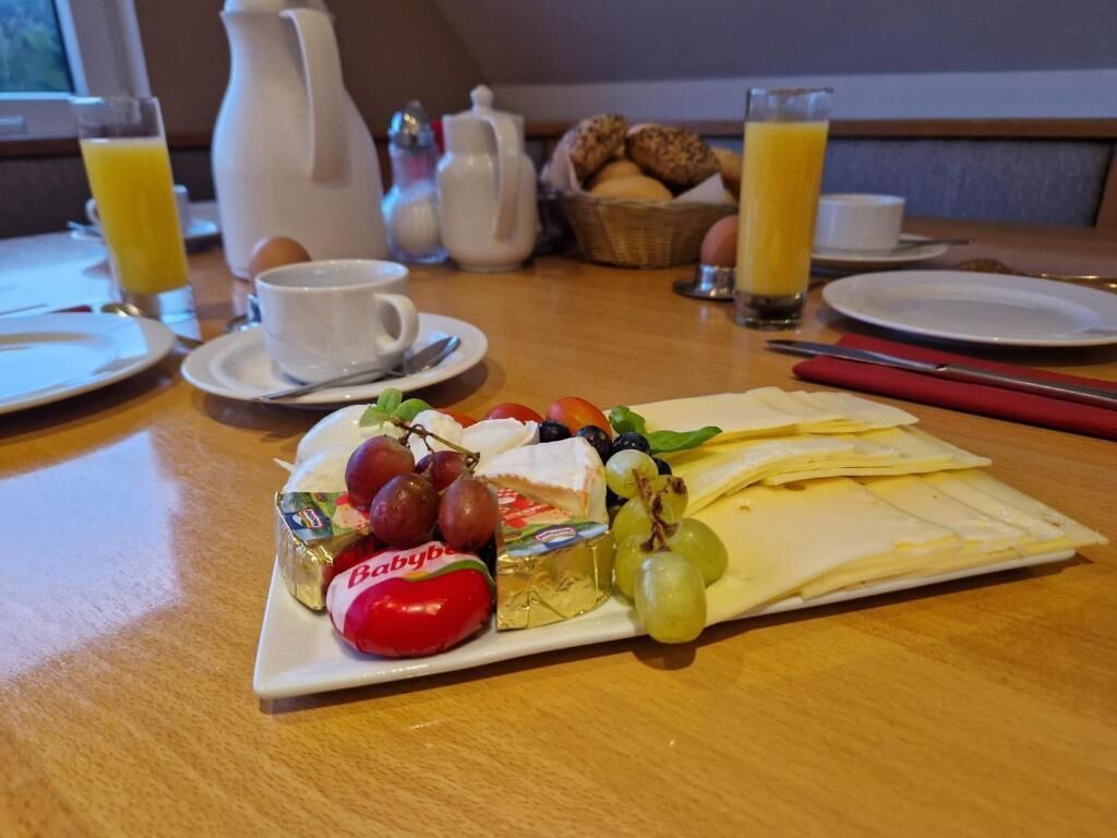 A breakfast table with a plate of cheese, grapes, and a tomato, along with a pitcher, a basket of bread, and glasses of orange juice.