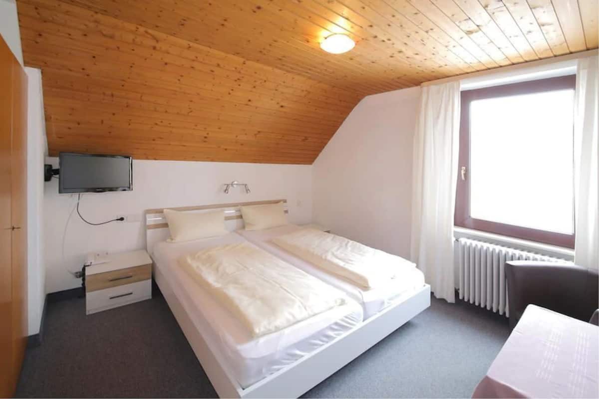 A bedroom with a wooden ceiling, a bed with white bedding, a bedside table, a TV mounted on the wall, and a window with curtains.