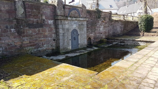 Un muro de piedra histórico con una reja metálica, un estrecho canal de agua y un camino empedrado.