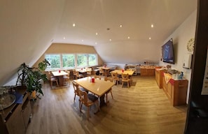 A spacious dining area with multiple tables and chairs, a kitchen area with wooden cabinets, and a large window providing natural light. - Eventhof Platzhirsch (Gaggenau)