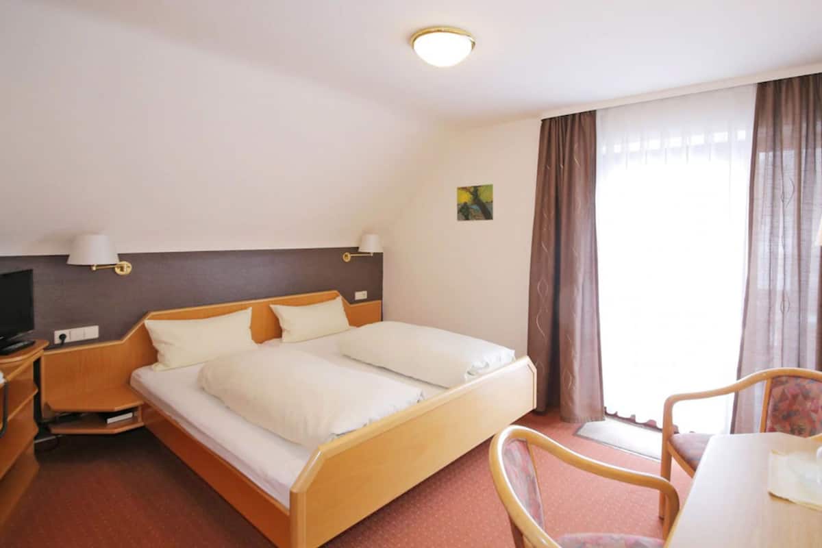A hotel room with a double bed, a bedside table with a lamp, a TV, a small table with a chair, and a window with curtains.