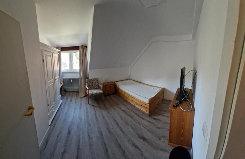 Une petite pièce étroite avec un plancher en bois, un lit simple, une chaise et un radiateur.