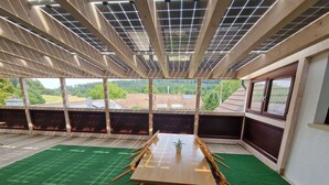 Una habitación con una mesa y sillas de madera, alfombra verde y ventanas amplias con vistas a edificios y árboles.