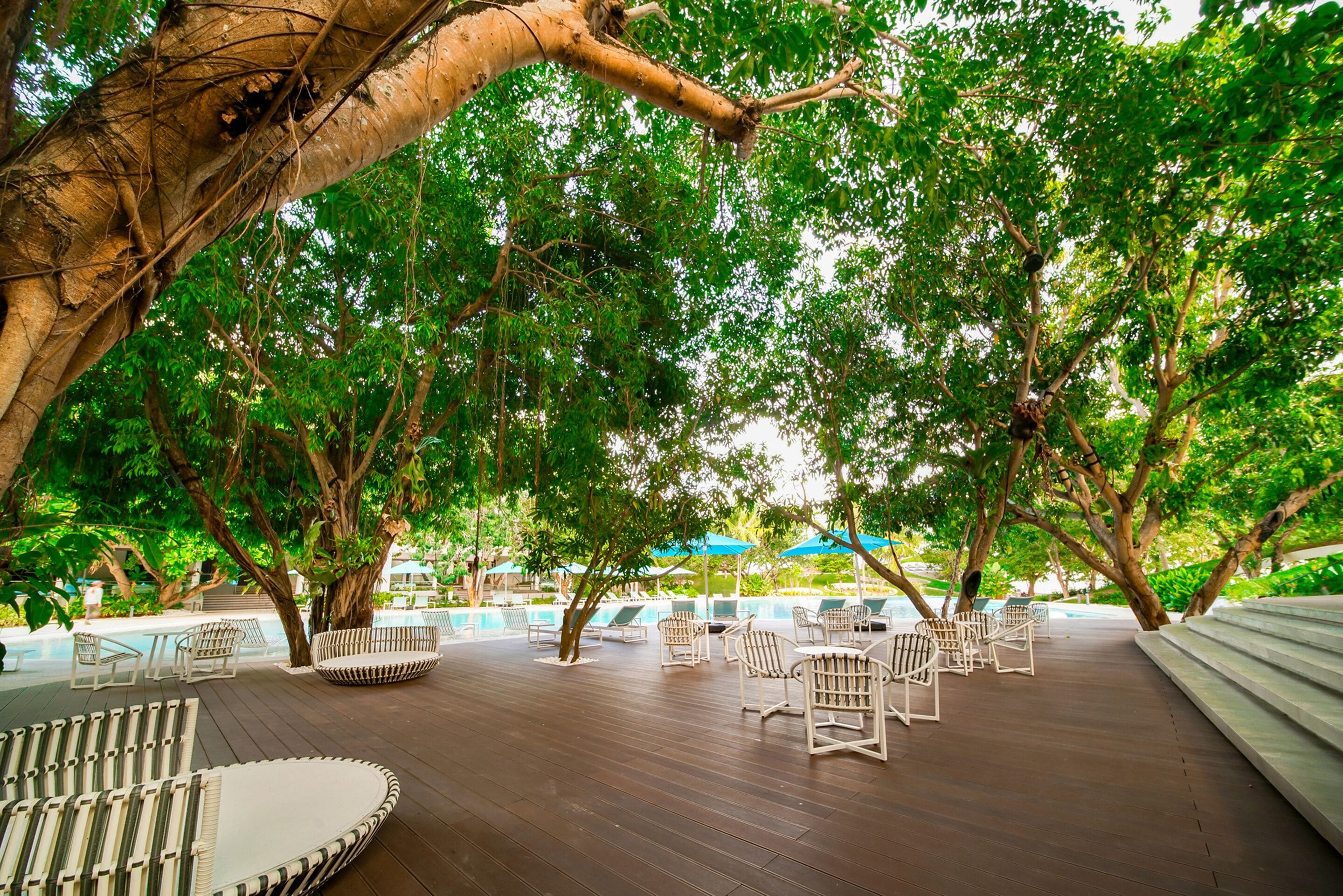 An outdoor lounge area with wooden flooring, white wicker furniture, and a view of a swimming pool.