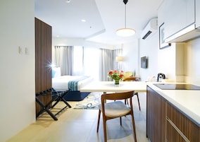 A modern hotel room with a bed, a dining area with a table and chairs, a kitchen area, and a bathroom.