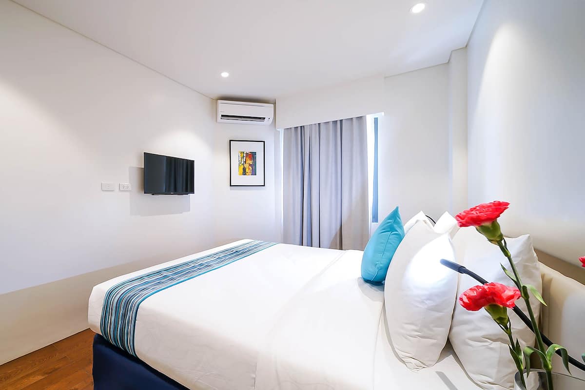 A modern hotel room with a neatly made bed, a mounted flat-screen TV, an air conditioning unit, and a window with curtains.
