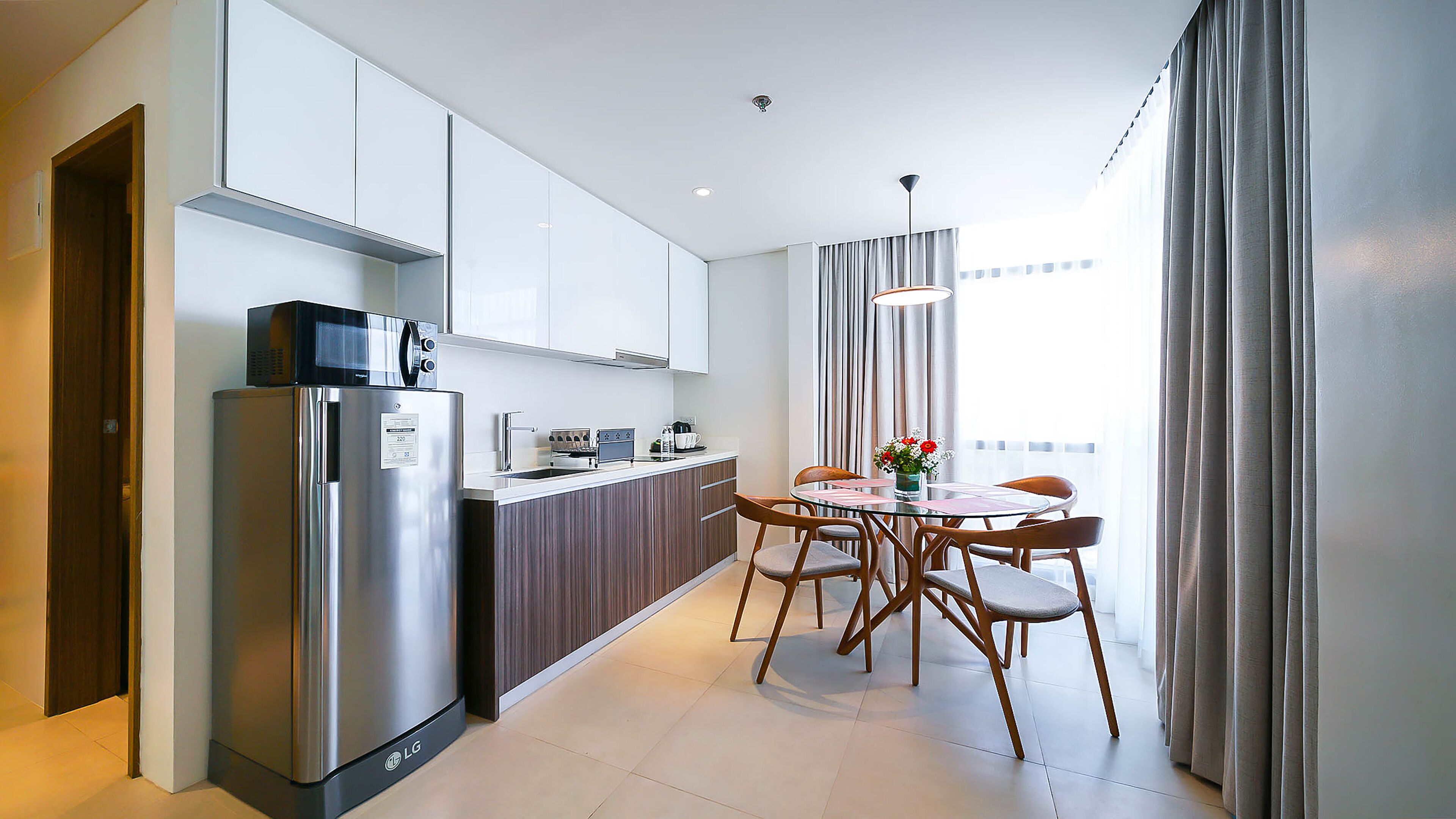 Una cocina moderna con armarios blancos, un refrigerador de acero inoxidable y un comedor con mesa de vidrio y sillas de madera.