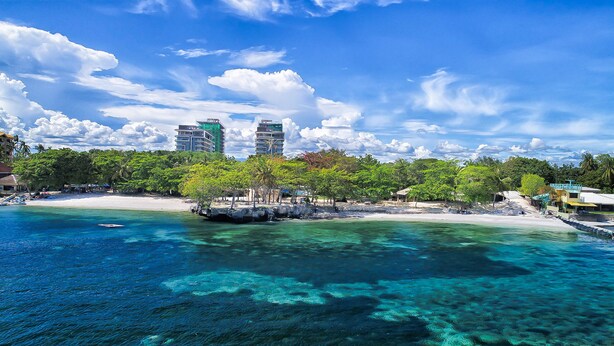 A coastal resort with clear turquoise waters, a sandy beach, and a backdrop of modern buildings. - Tambuli Seaside Resort and Spa (Lapu-Lapu)