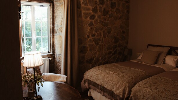 Un dormitorio con dos camas, una pared de piedra, una ventana con cortinas, una lámpara y una mesita con una planta.