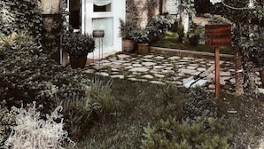 Una casa de piedra con puerta y ventanas blancas, rodeada de abundante vegetación y un camino de piedra.