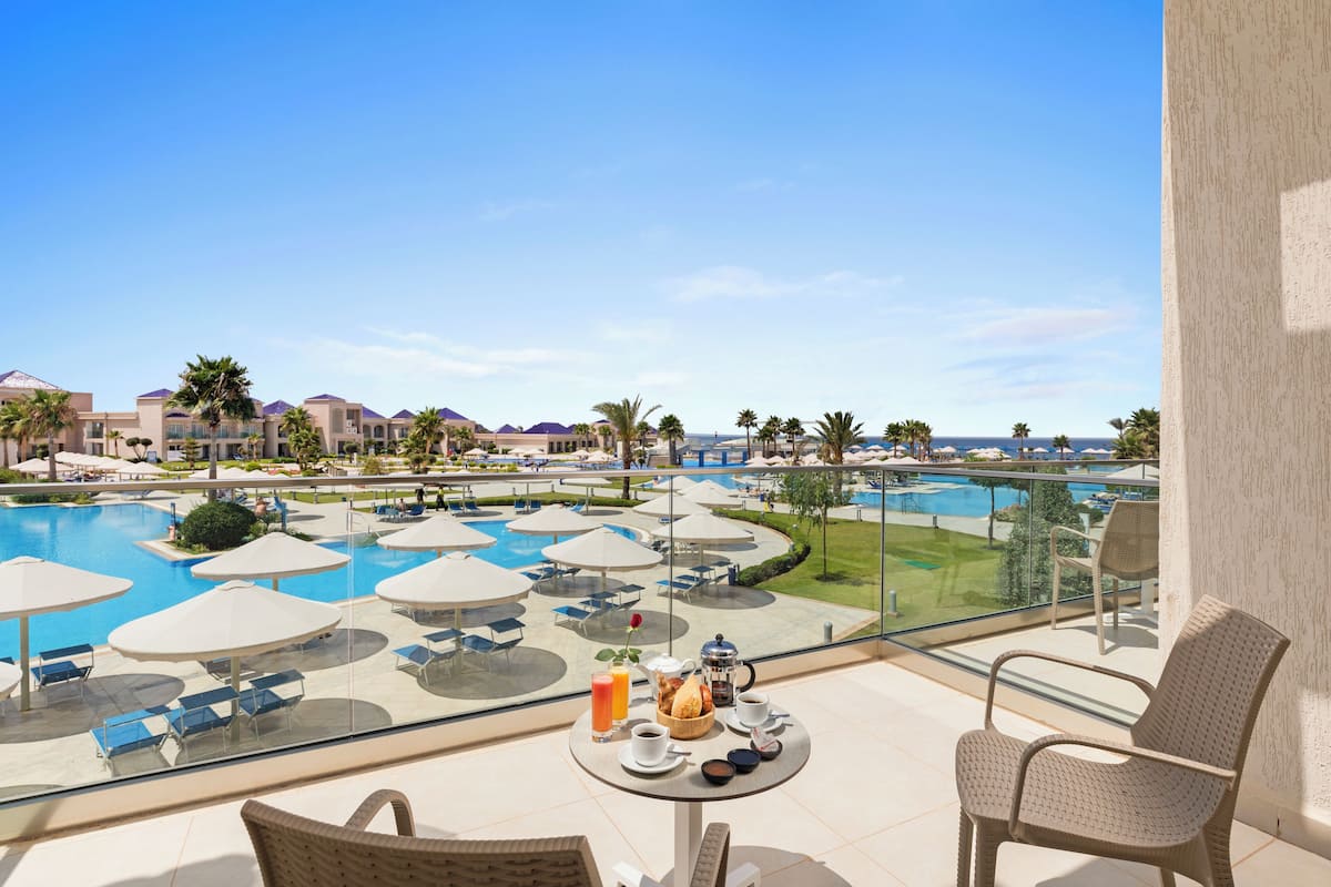 A balcony with a view of a swimming pool, lounge chairs, and a table with breakfast items.