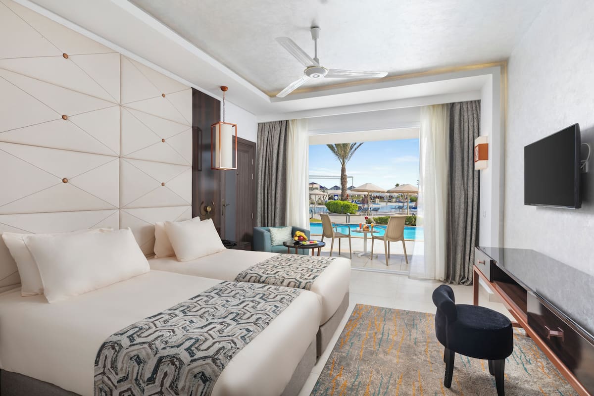 A hotel room with a large bed, a TV, a desk, and a view of a pool and palm trees.