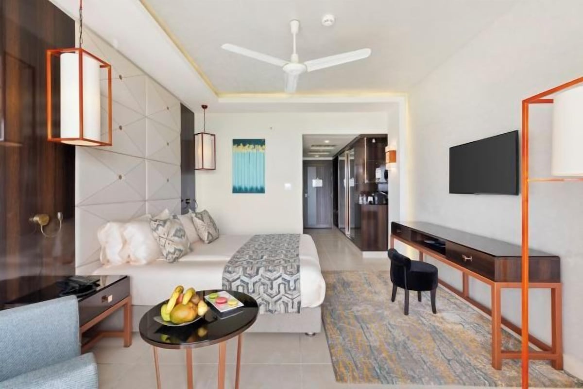 Una habitación de hotel moderna con cama, escritorio, silla, televisor y mesa de comedor con frutas.