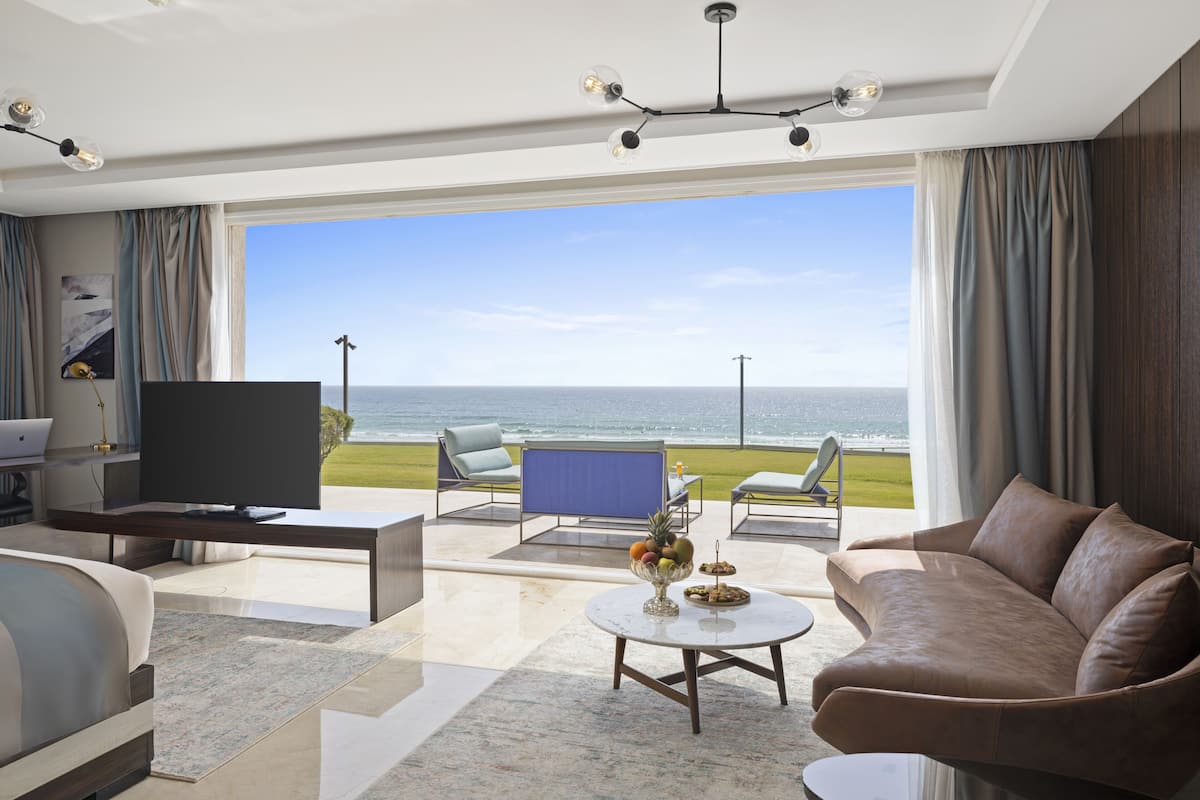 A modern living room with a large TV, comfortable seating, a coffee table with fruit, and a view of the ocean.