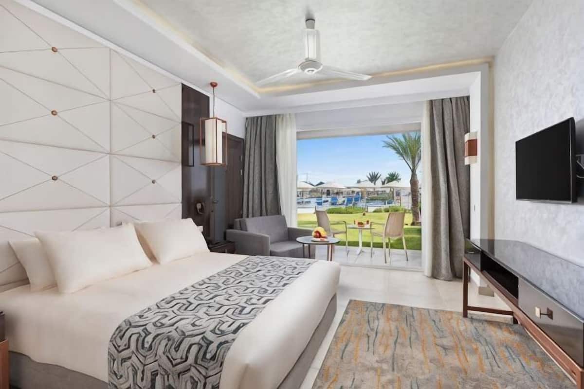 A hotel room with a bed, a TV, a ceiling fan, and a view of a pool and palm trees.
