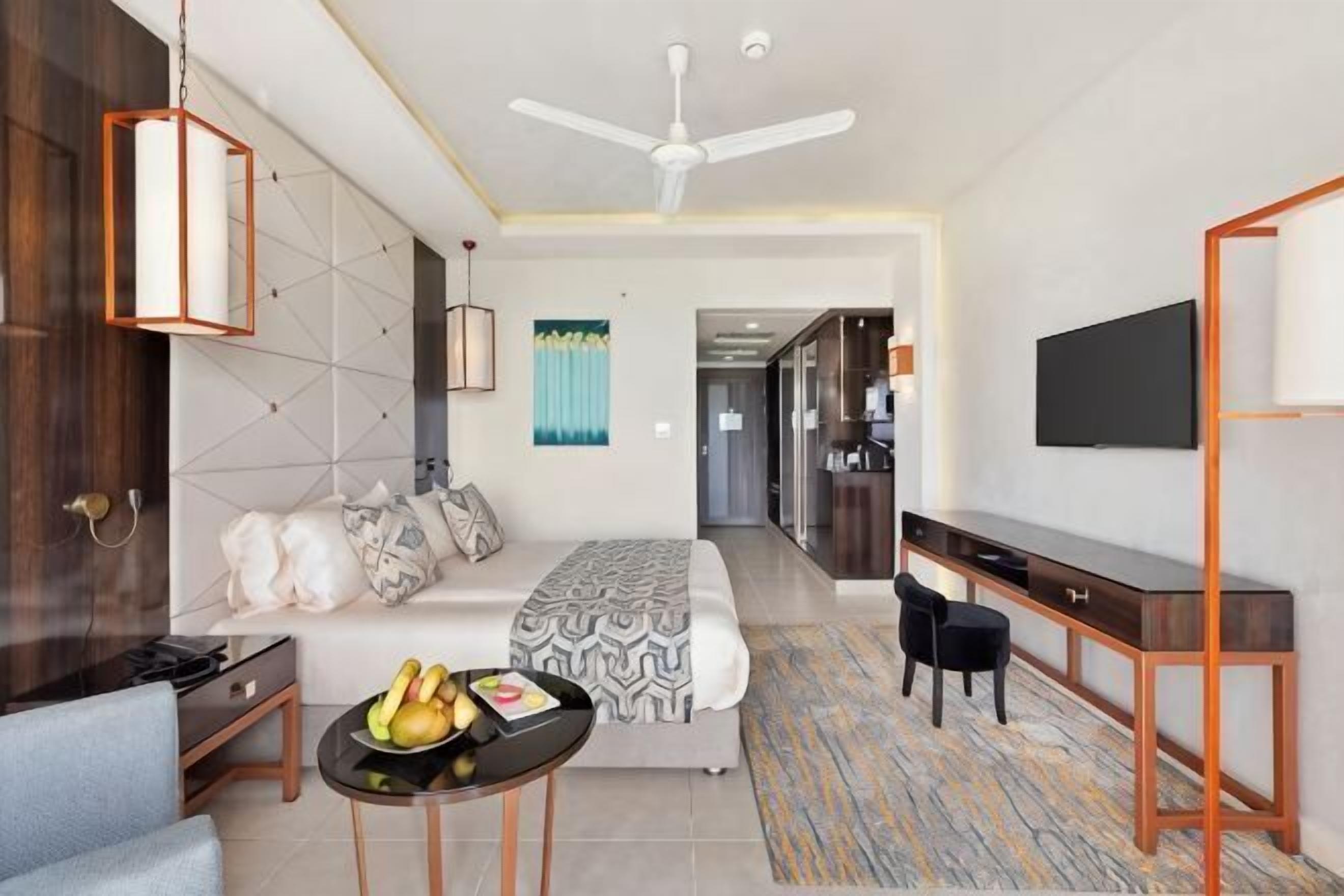 A modern hotel room with a bed, sofa, dining table, TV, and a ceiling fan.