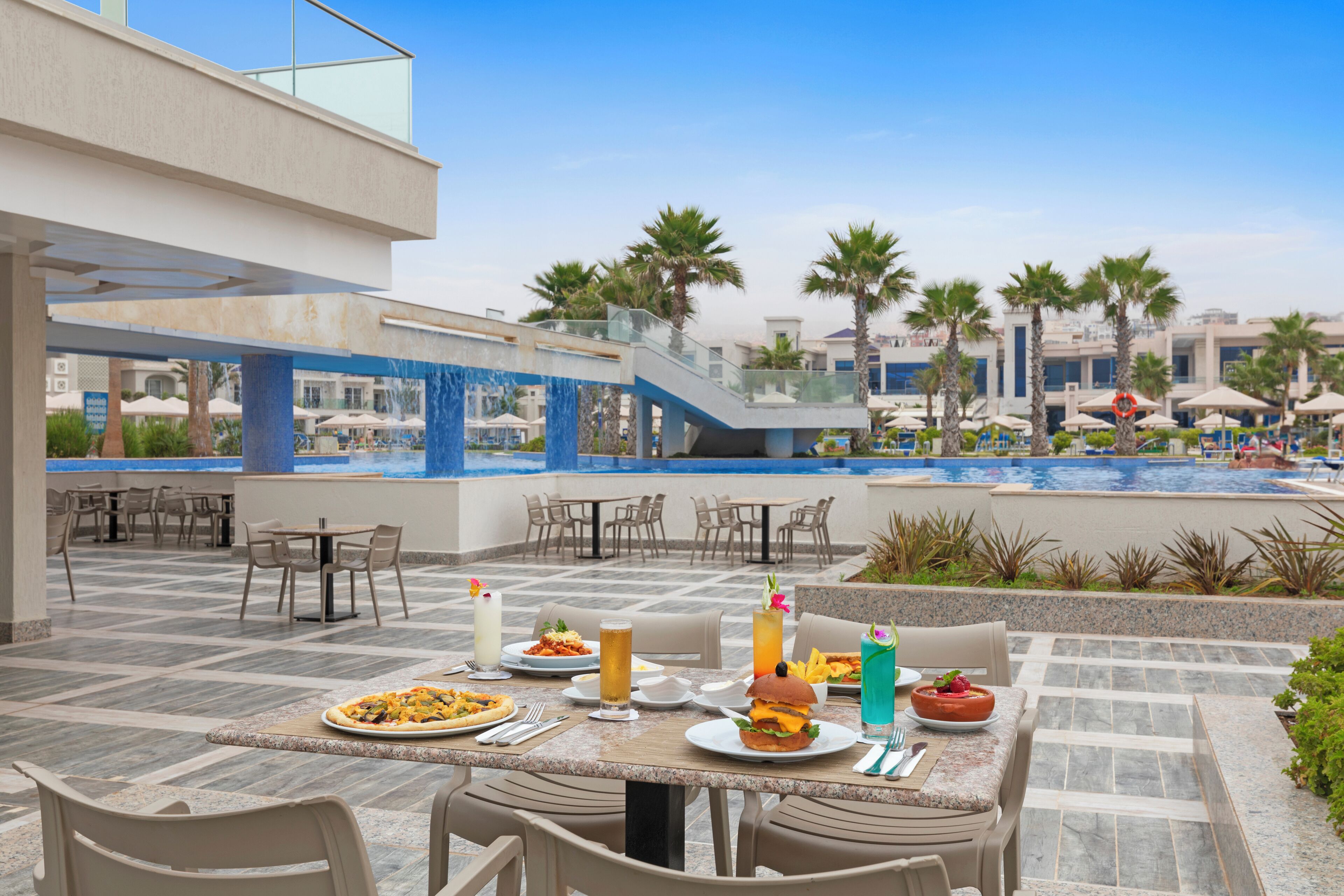 Un espacio exterior para comer con mesas y sillas, una piscina y palmeras al fondo.