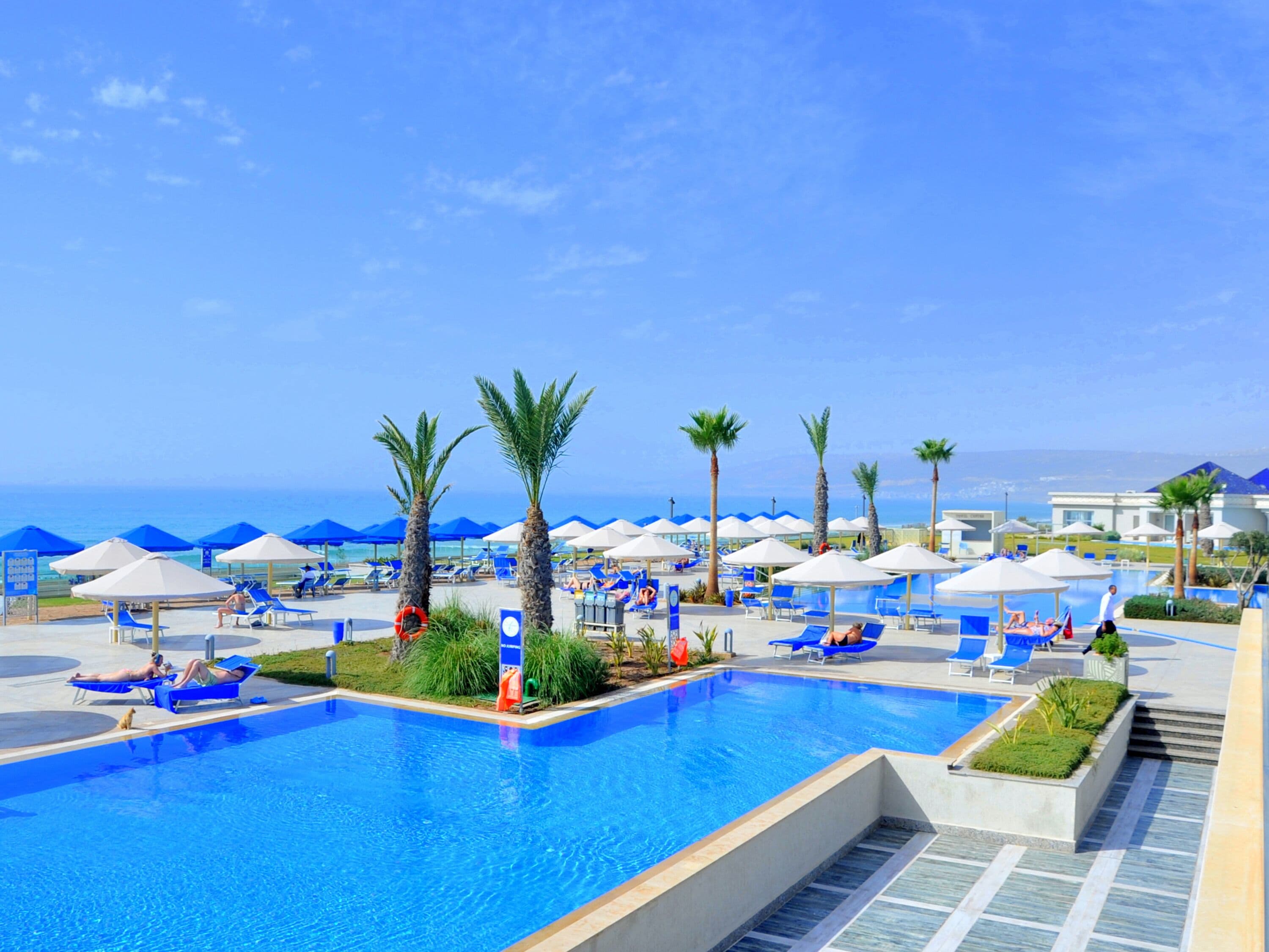 A pool area with blue lounge chairs and umbrellas, palm trees, and a clear view of the ocean.