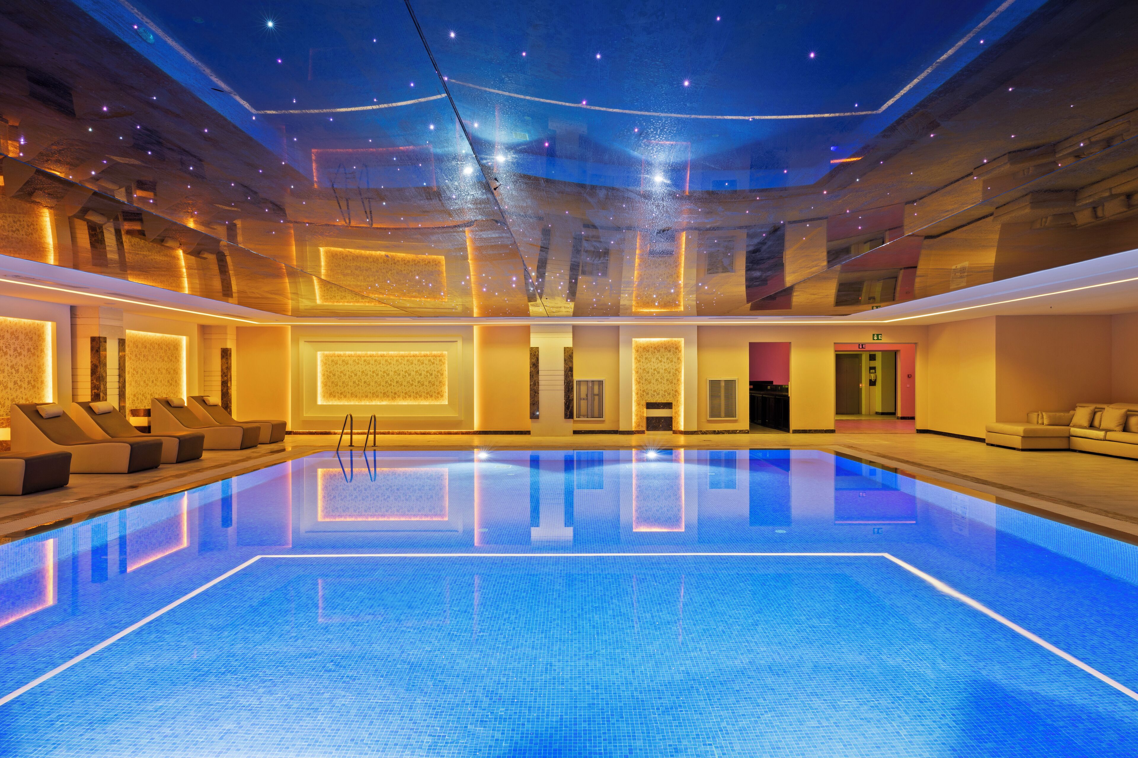 A large indoor swimming pool with a tiled floor and walls, surrounded by lounge areas with seating.