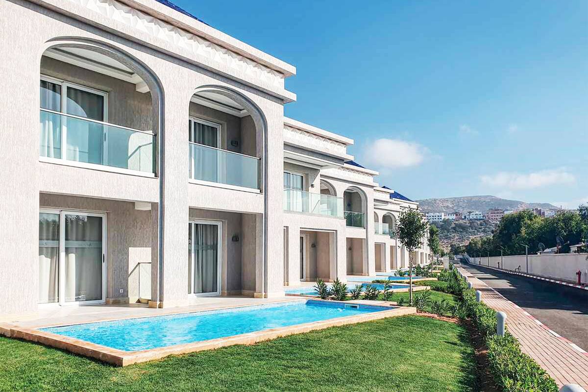 A modern hotel with a swimming pool, multiple balconies, and a view of the city in the background.