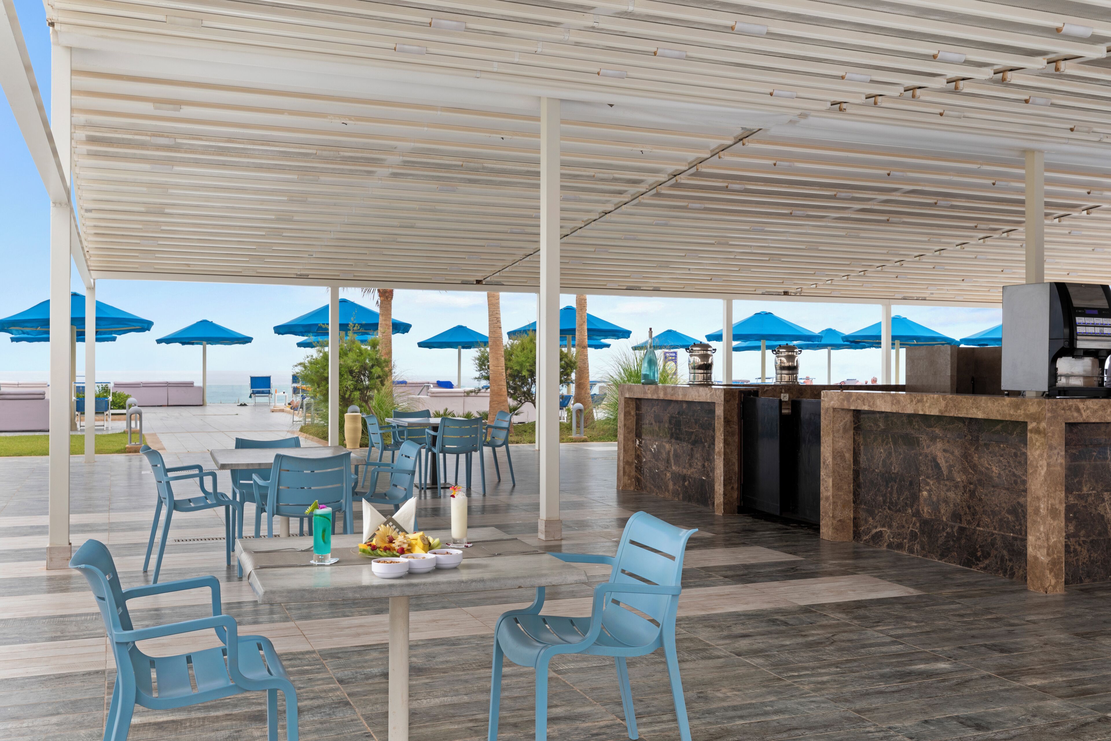 Un espace extérieur aménagé avec des chaises et des parasols bleus, un comptoir où est affiché le menu, et une vue sur la mer.
