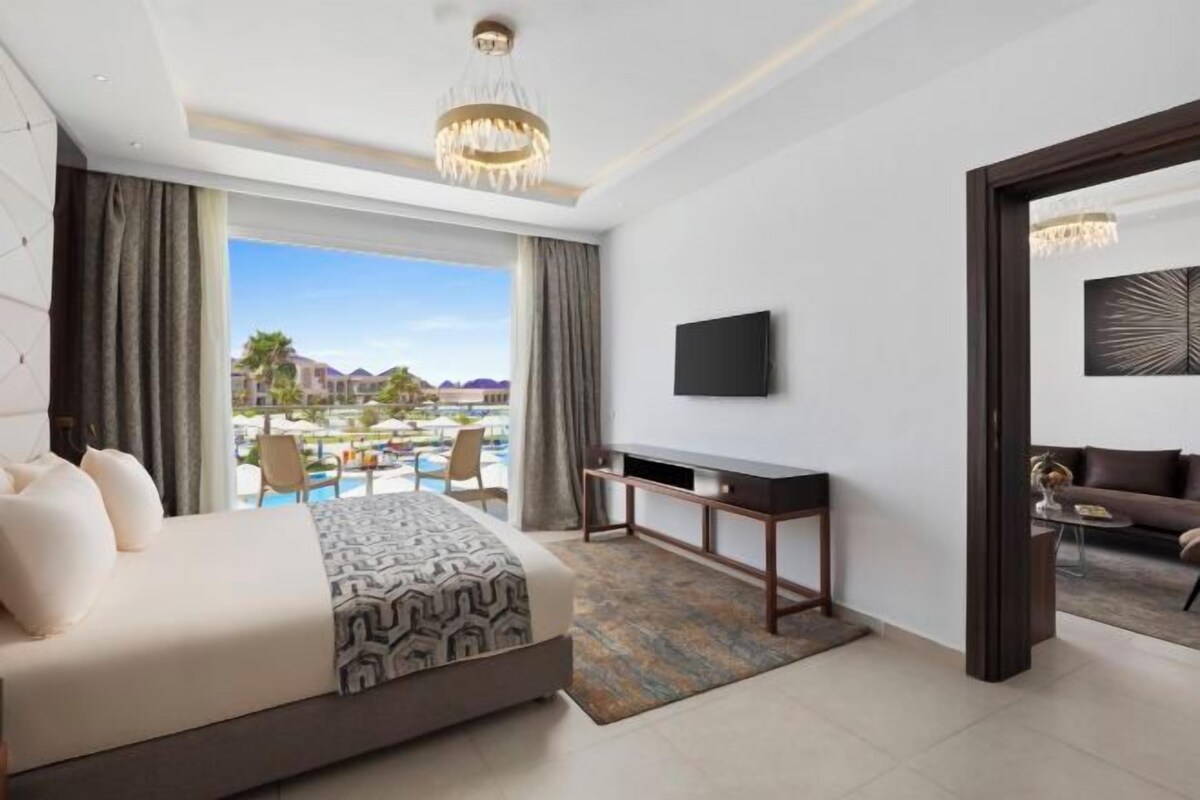 A hotel room with a bed, a desk, a TV, and a view of a pool and buildings.