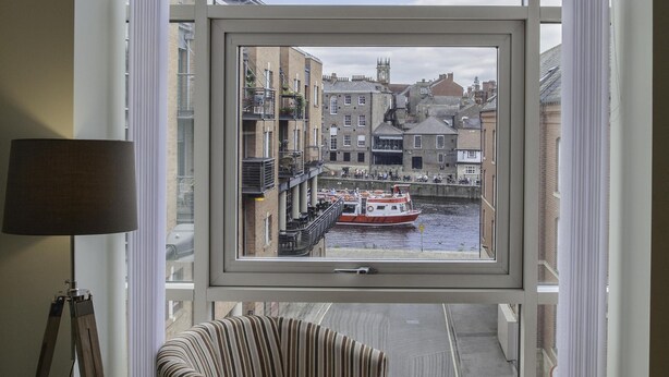 Une pièce dotée d’une grande fenêtre donnant sur un canal où naviguent des bateaux, avec des fauteuils à rayures, un petit pouf et un lampadaire.