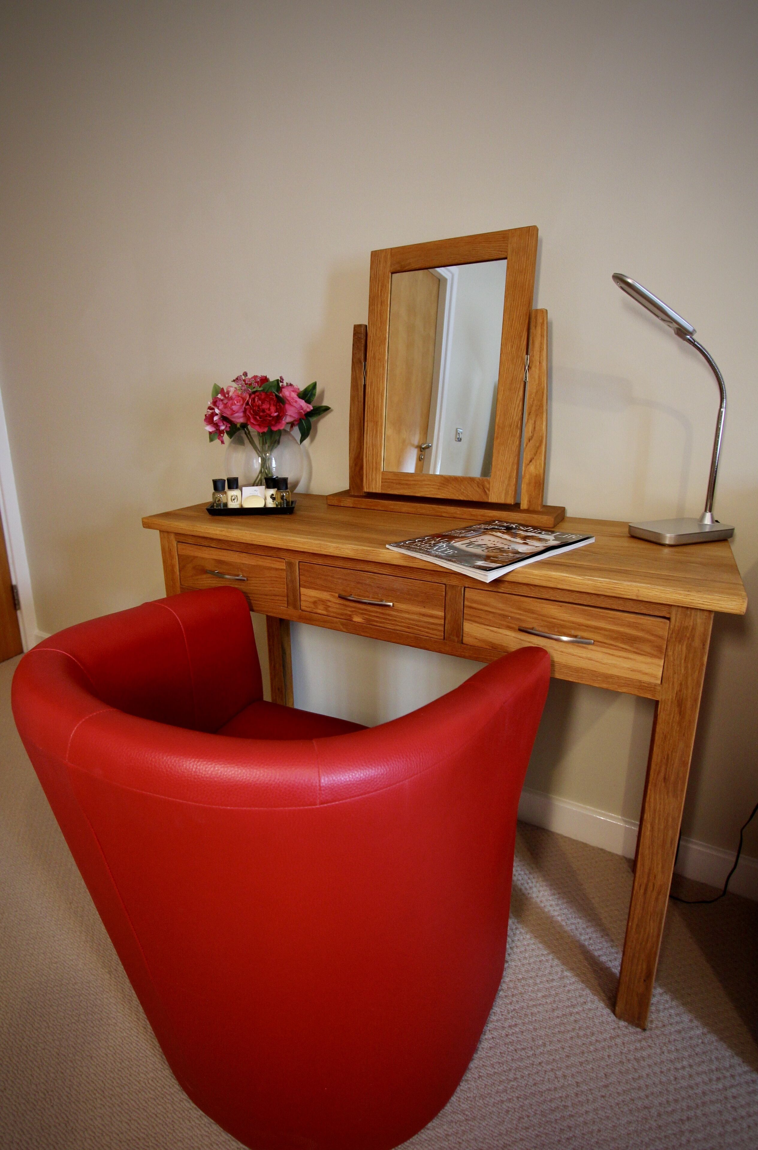 A wooden desk with a mirror, a red chair, a vase with flowers, and a magazine.